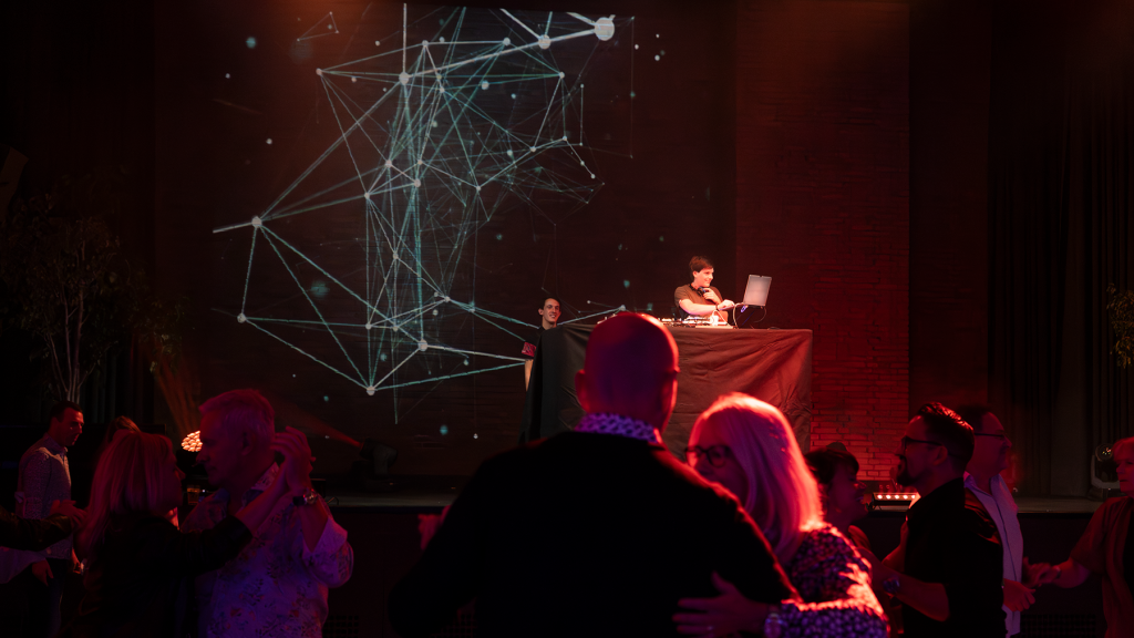 Menschen tanzen vor einer Bühne, auf der eine Person an einem DJ-Pult Musik auflegt. Im Hintergrund ist eine große Leinwand mit abstrakten, vernetzten Linien und Punkten in heller Farbe auf dunklem Hintergrund zu sehen. Die Szene ist in warmes, rotes Licht getaucht.