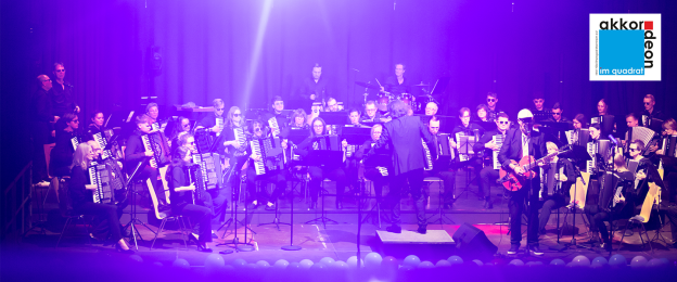 Auf einer Bühne spielt ein großes Akkordeonorchester vor Publikum. Die Musikerinnen und Musiker sitzen in mehreren Reihen und tragen überwiegend dunkle Kleidung. Im Mittelpunkt steht eine dirigierende Person vor dem Ensemble. Rechts auf der Bühne tritt zusätzlich eine Person mit E-Gitarre auf. Die Bühne ist in violettes und pinkfarbenes Licht getaucht. Im Hintergrund sind Schlagzeug und weitere Instrumente zu sehen. Oben rechts befindet sich das Logo „akkordeon im quadrat“.