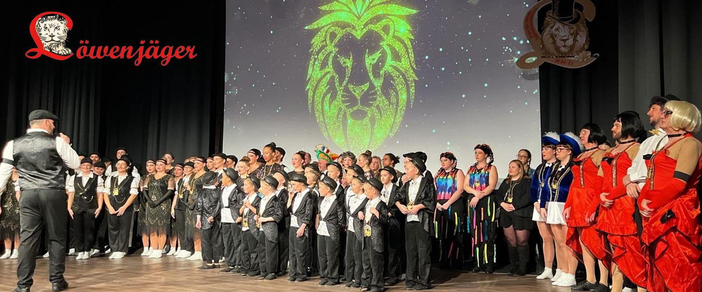 Bühnenszene mit einer großen Gruppe von Mitwirkenden unterschiedlichen Alters. Die Personen stehen in mehreren Reihen auf einer Bühne und tragen verschiedene Kostüme, darunter schwarze, rote, blaue und mehrfarbige Outfits. Im linken Vordergrund steht eine Person mit dem Rücken zum Publikum und spricht in ein Mikrofon. Im Hintergrund wird auf einer Leinwand ein großer stilisierter grüner Löwenkopf projiziert. Im oberen Bildbereich ist der Schriftzug „Löwenjäger“ zu sehen. Die Aufnahme zeigt einen gemeinsamen Bühnenauftritt oder Abschlussmoment einer Veranstaltung.